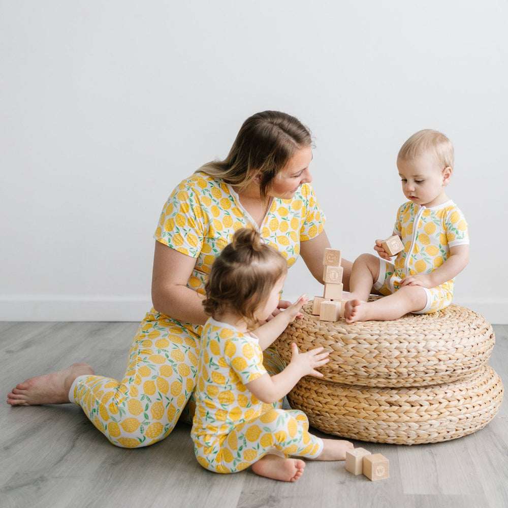 Image of mom and two kids playing with wooden building blocks. They are all wearing matching short sleeve pajamas in Lemons print. This print features vibrant pops of yellow fruit with green accents that sit upon a white background with white trim.