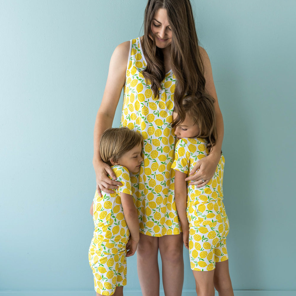 Image of mom and her two kids, all wearing matching Lemons printed pajamas that are perfect for lounging around this summer! This Lemons print is sure to brighten your day! This signature print features vibrant pops of yellow fruit with green accents that sit upon a white background with white trim.