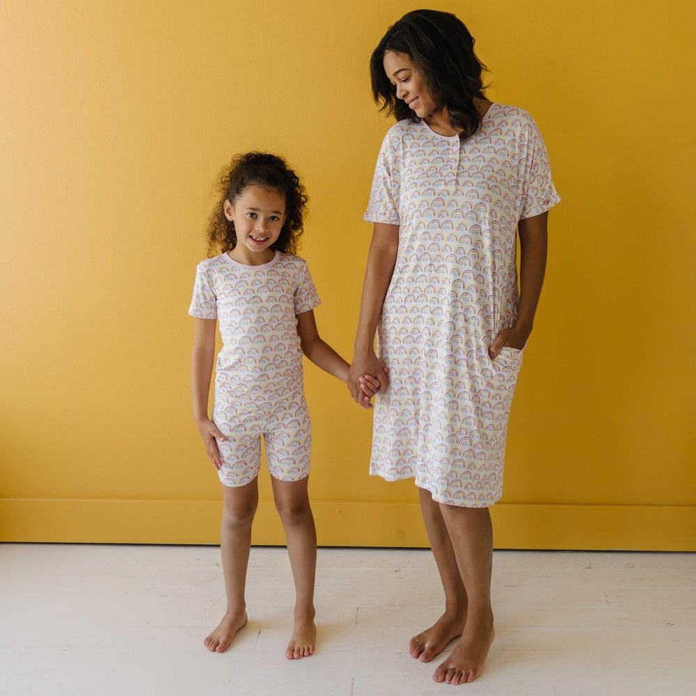 Image of mom and daughter wearing matching rainbow printed pajamas. The daughter is shown wearing a short sleeve and shorts pajama set and the mom is shown wearing a caftan gown. This print features multicolored rainbows that sit upon a white background w