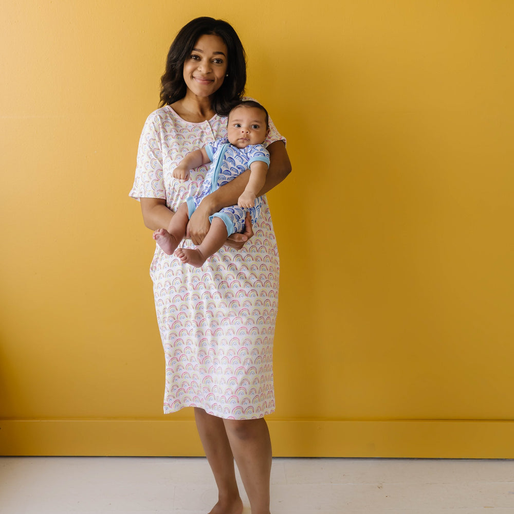 Click to see full screen - Image of mom holding her infant son. They are both shown wearing coordinating rainbow printed pajamas, with the mom shown wearing a caftan gown, and the infant son shown wearing a short sleeve and shorts zip up romper.