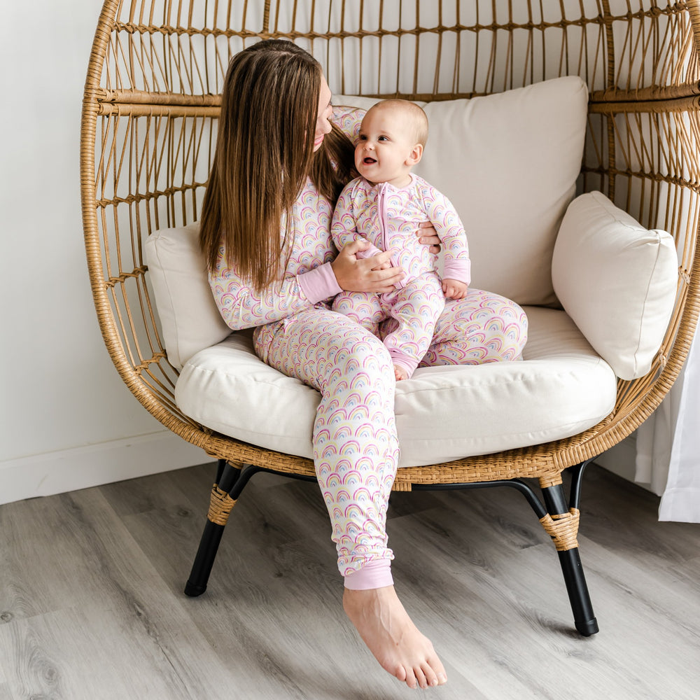Image of mom and her daughter wearing matching rainbow printed pajamas. This print features multicolored rainbows that sit upon a white background with light pink accent trim.
