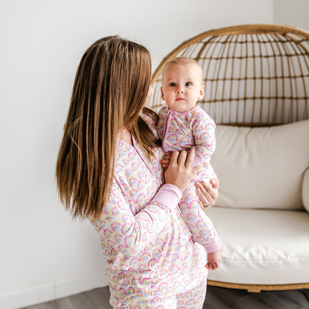 Image of mom and her daughter wearing matching rainbow printed pajamas. This print features multicolored rainbows that sit upon a white background with light pink accent trim.