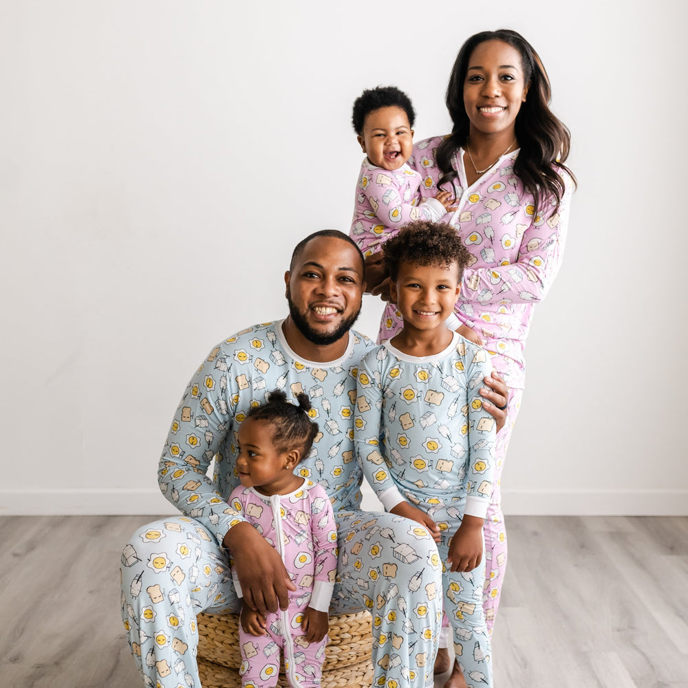 Click to see full screen - Image of family of five all wearing matching Breakfast Buddies printed pajamas. Father and son are wearing light Blue Breakfast Buddies printed pajamas with white trim accents, while mom is shown wearing Pink Breakfast Buddies printed pajamas with white trim accents. The two daughters in the photo are also wearing light pink breakfast printed zip up rompers with white trim accents. The breakfast foods featured on this print include sunny side up eggs, toast, and milk.