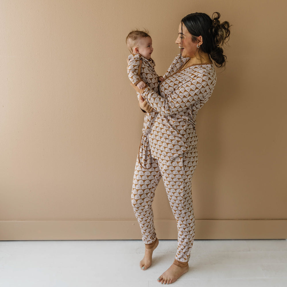 Click to see full screen - Image of mom and infant son wearing matching pajamas in Rust Rainbows print. The mom is shown wearing a long sleeve pajama top with coordinating pants, while her son is shown in an infant knotted gown. This print features white rainbows that sit upon a rust brown background with matching rust brown trim.