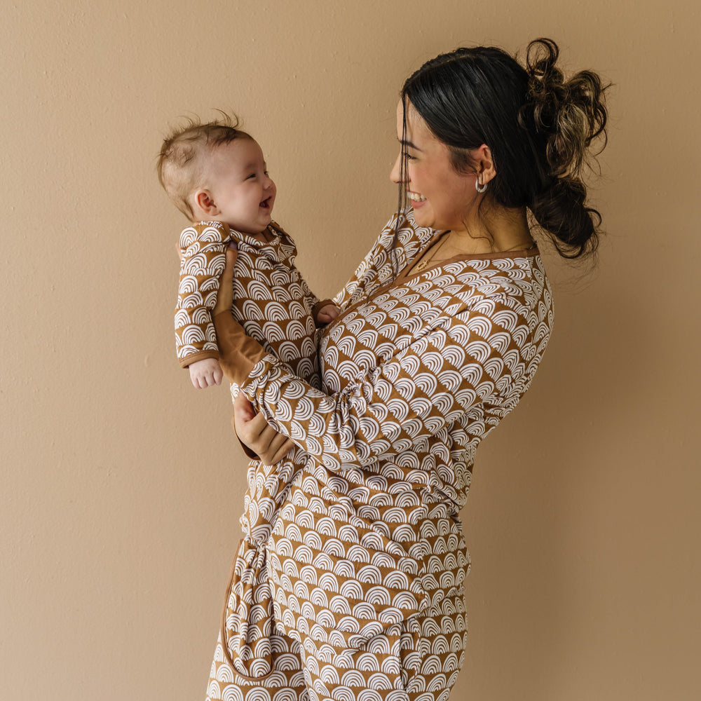 Image of mom and infant son wearing matching pajamas in Rust Rainbows print. The mom is shown wearing a long sleeve pajama top, while her son is shown in an infant knotted gown. This print features white rainbows that sit upon a rust brown background with matching rust brown trim.