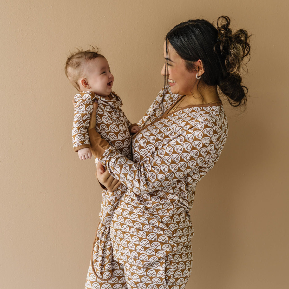 Image of mom and infant son wearing matching pajamas in Rust Rainbows print. The mom is shown wearing a long sleeve pajama top, while her son is shown in an infant knotted gown. This print features white rainbows that sit upon a rust brown background with matching rust brown trim.