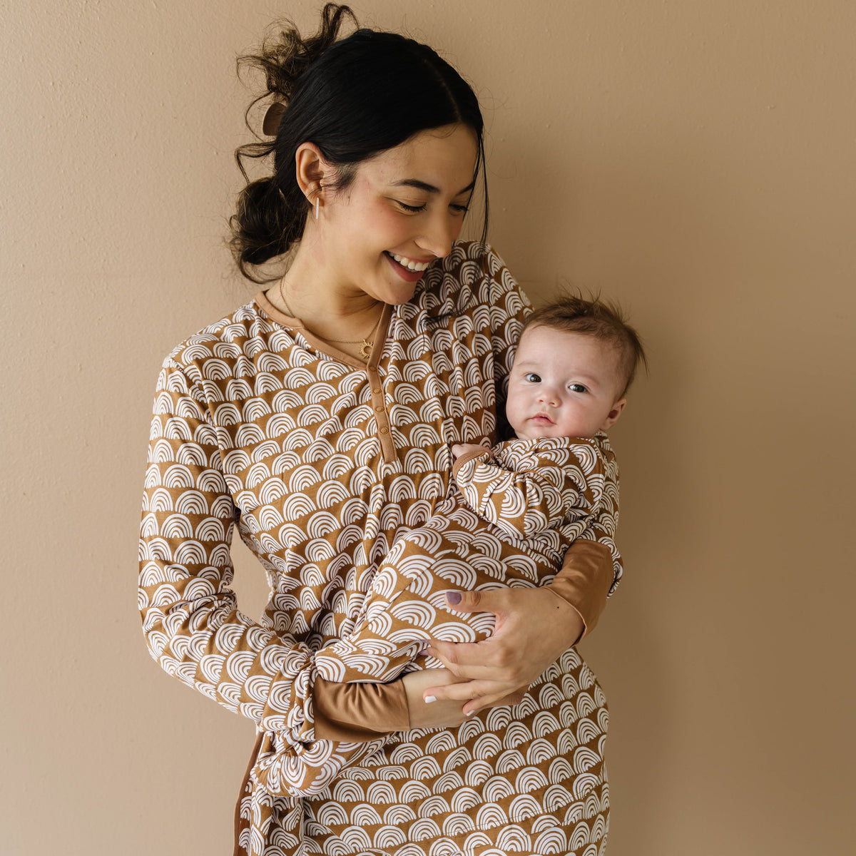 Image of mom and infant son wearing matching pajamas in Rust Rainbows print. The mom is shown wearing a long sleeve pajama top, while her son is shown in an infant knotted gown. This print features white rainbows that sit upon a rust brown background with matching rust brown trim.