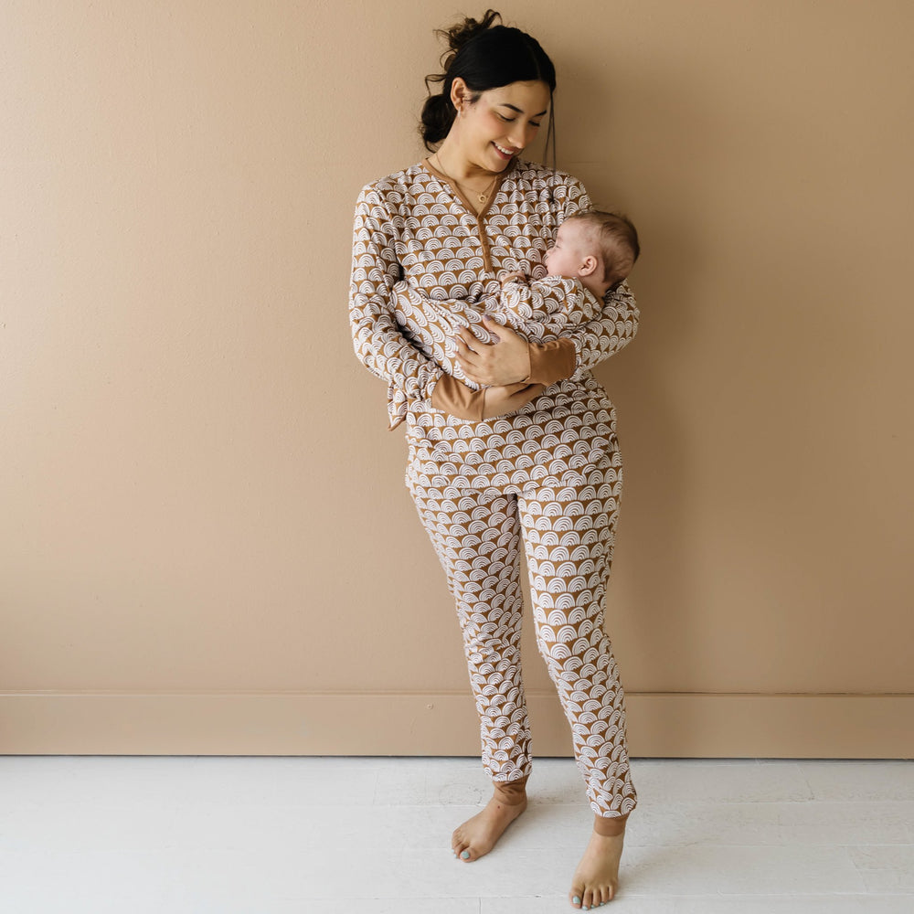 Image of mom and infant son wearing matching pajamas in Rust Rainbows print. The mom is shown wearing a long sleeve pajama top and coordinating pajama pants, while her son is shown in an infant knotted gown. This print features white rainbows that sit upon a rust brown background with matching rust brown trim.
