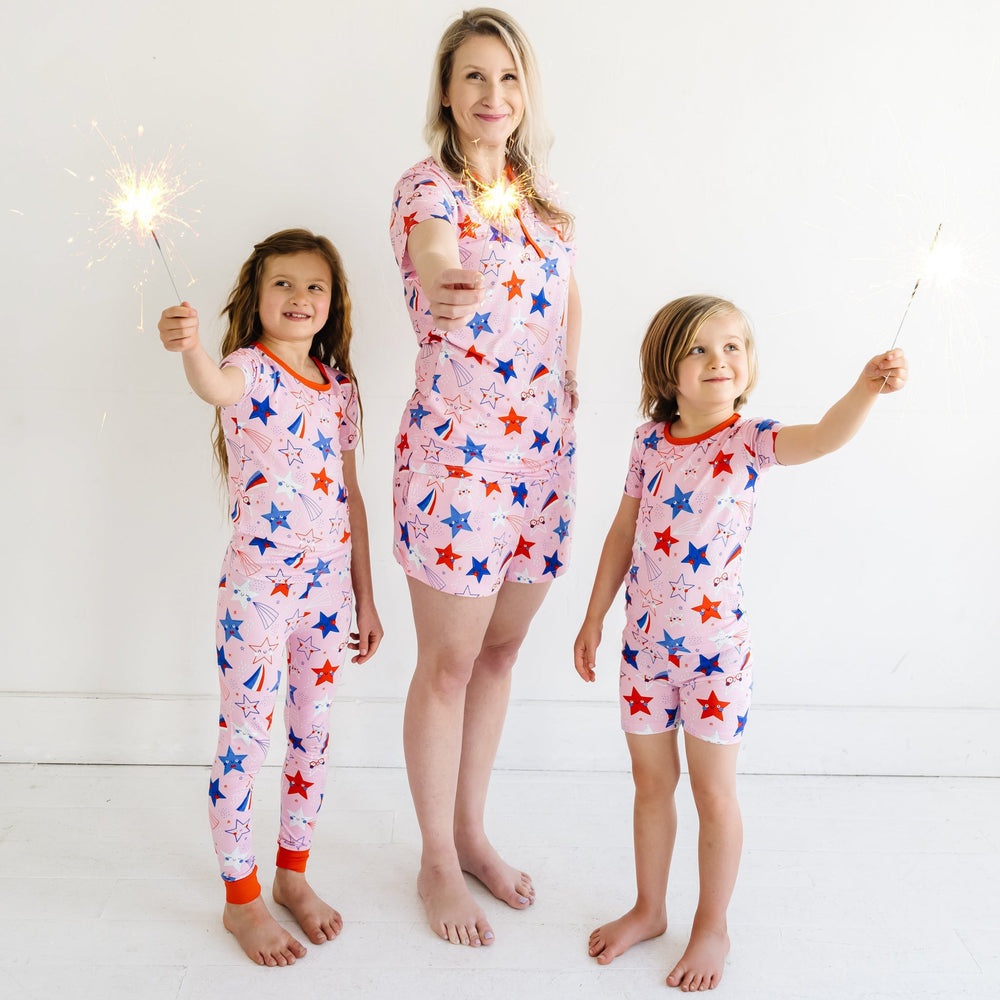 Mother and her two children playing with sparklers wearing matching Pink Stars and Stripes printed pajama sets. Mother is wearing a Pink Stars and Stripes printed women's pajama top and matching women's pajama shorts. One child is wearing Pink Stars and Stripes printed two piece short sleeve and shorts pajama set and the other is wearing a matching short sleeve pajama set
