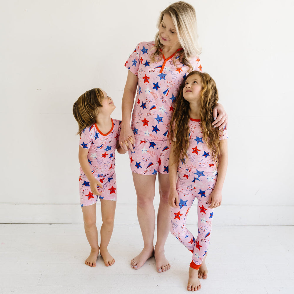Mother and her two children wearing matching Pink Stars and Stripes printed pajama sets. Mother is wearing a Pink Stars and Stripes printed women's pajama top and matching women's pajama shorts. One child is wearing Pink Stars and Stripes printed two piece short sleeve and shorts pajama set and the other is wearing a matching short sleeve pajama set