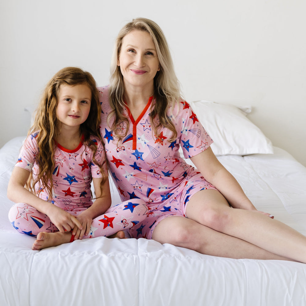 Mother and her daughter sitting together on a bed wearing matching Pink Stars and Stripes printed pajama sets. Mother is wearing a Pink Stars and Stripes printed women's pajama top and matching women's pajama shorts. Child is wearing Pink Stars and Stripes printed two piece short sleeve pajama set