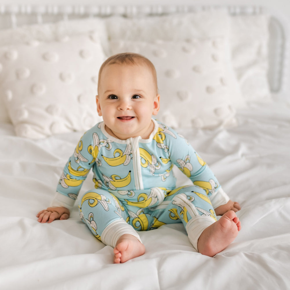 Baby boy wearing banana printed zip up romper. This banana printed zip up romper features a light blue background with pops of yellow and white trim accents on the collar, zip feature, sleeve cuffs, and pant cuffs. 