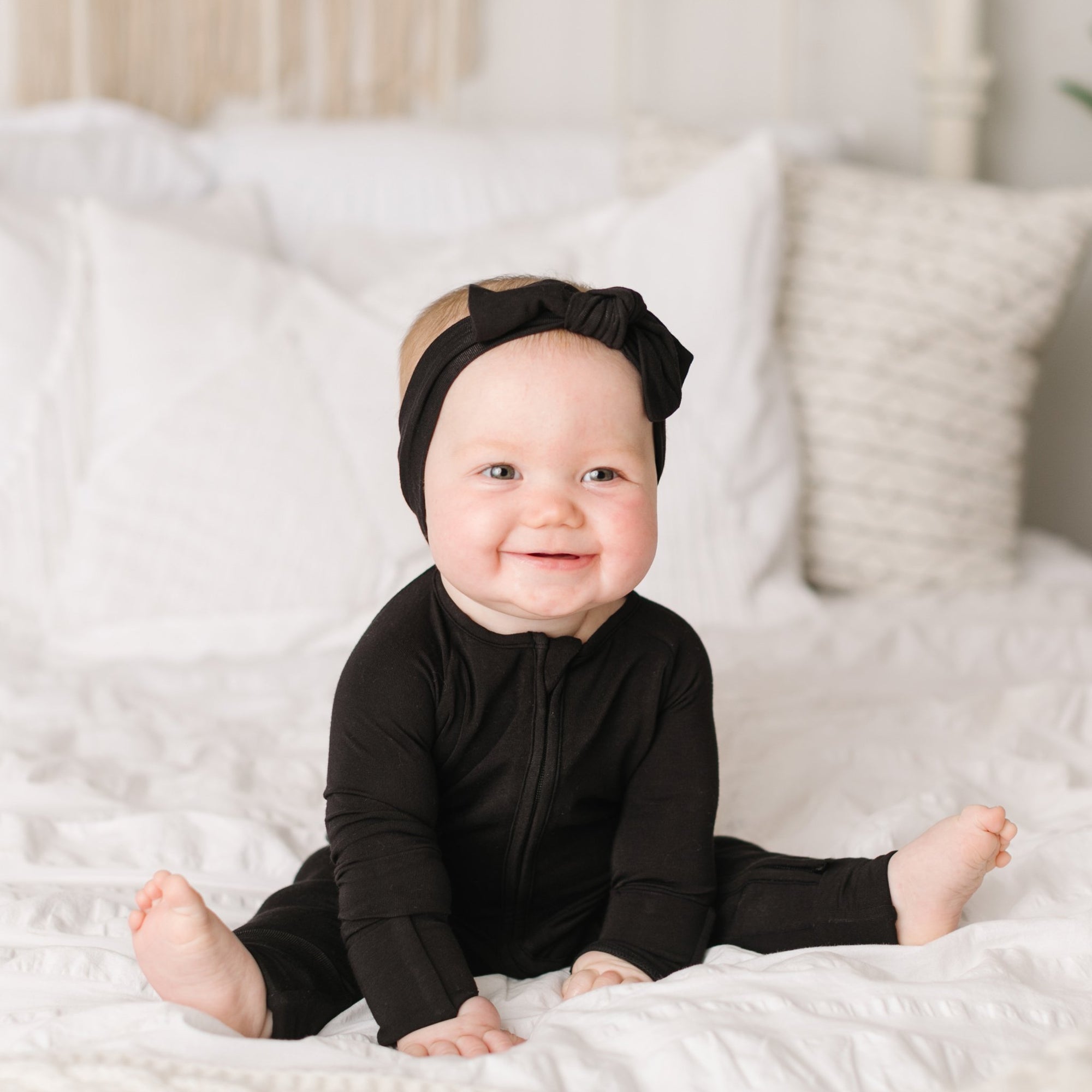 Image of baby girl sitting on a bed wearing solid black zip up romper with matching solid black bow headband. 
