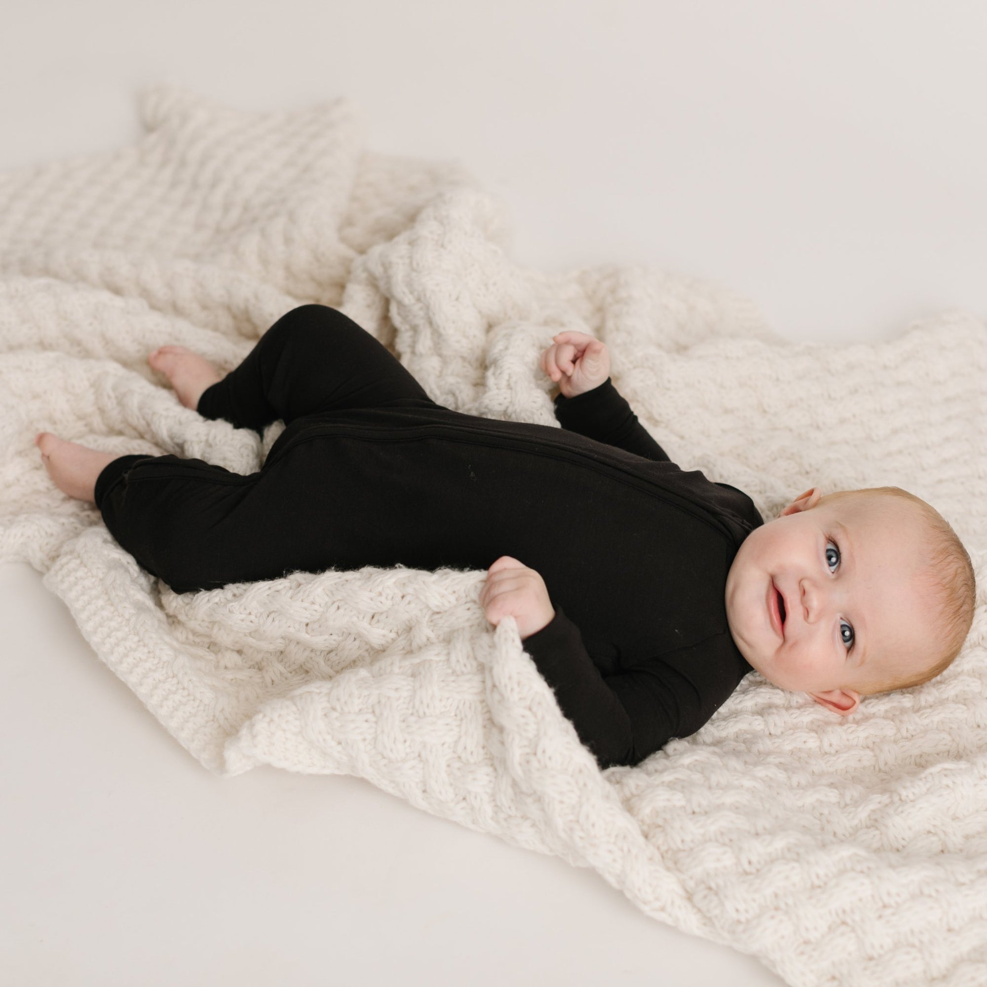 Image of baby boy laying down on a blanket wearing solid black zip up romper. 