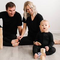 Image of family of 3 sitting on the floor and playing with wooden building blacks. They are all shown wearing matching solid black pajamas. The mom and dad are both shown wearing solid black short sleeve pj tops with matching solid black pj pants and the 