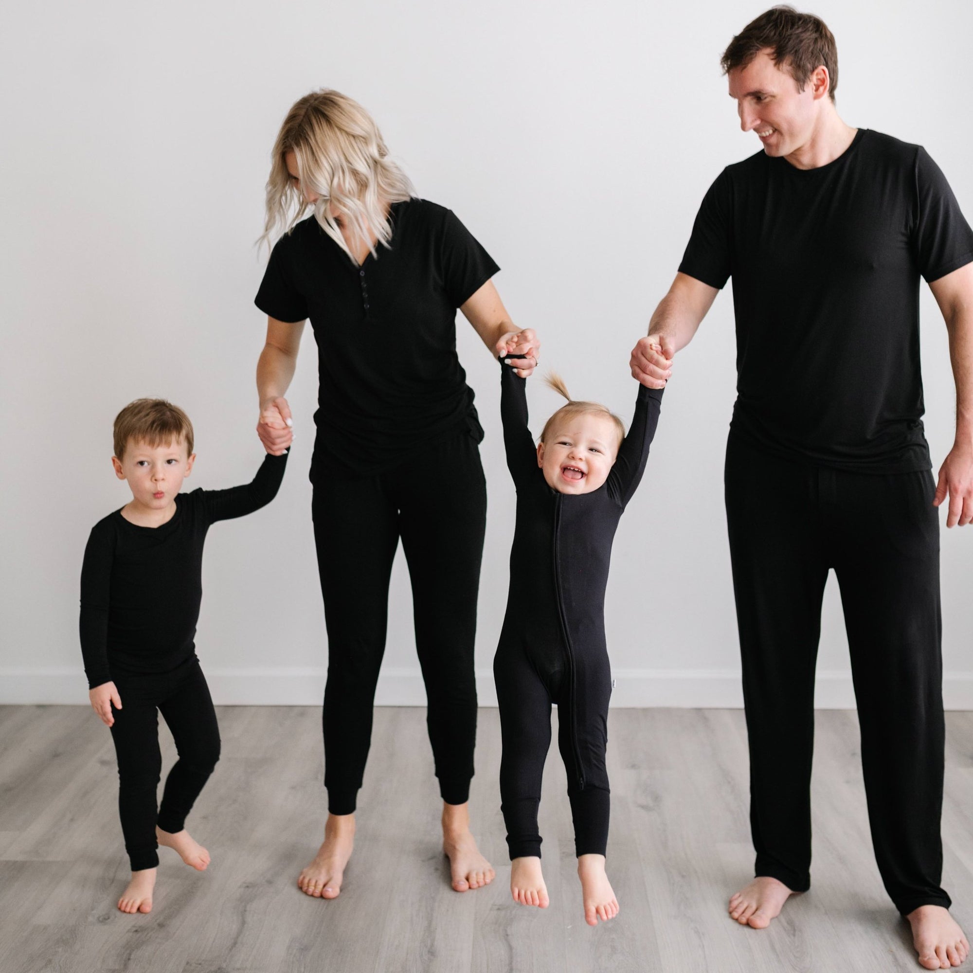 Image of family of 4 all holding hands. They are all wearing matching solid black pajamas. The mom and dad are both shown wearing solid black short sleeve pj tops with matching solid black pj pants, the son is shown wearing a long sleeve pajama set, and t