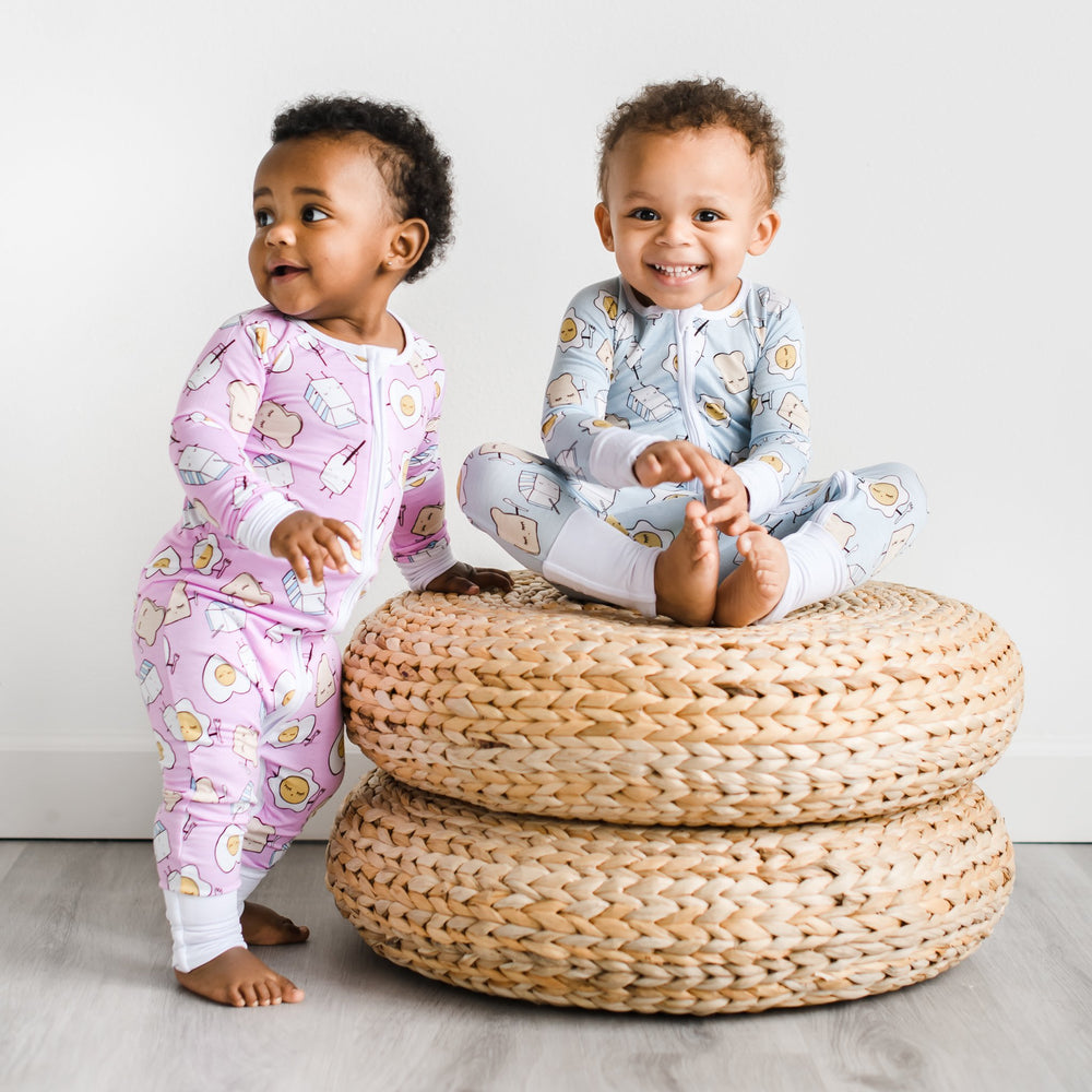 Click to see full screen - Image of baby boy and baby girl wearing Breakfast Buddies printed Zippies. This print has a light blue background with white trim accents and the breakfast foods featured on this print include sunny side up eggs, toast, and milk.