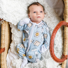 Image of baby boy wearing Blue Breakfast Buddies printed zip up Zippy. This print has a light blue background with white trim accents and the breakfast foods featured on this print include sunny side up eggs, toast, and milk.