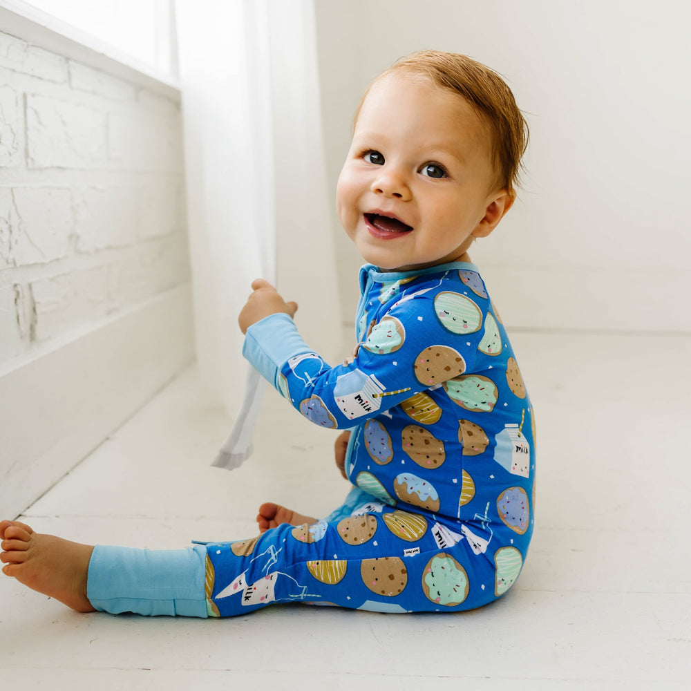 Image of toddler boy wearing a zip up romper in cookies and milk print. This print features milk cartons, colorful sprinkled cookies, and chocolate chip cookies that sit upon a blue background with sky blue trim.