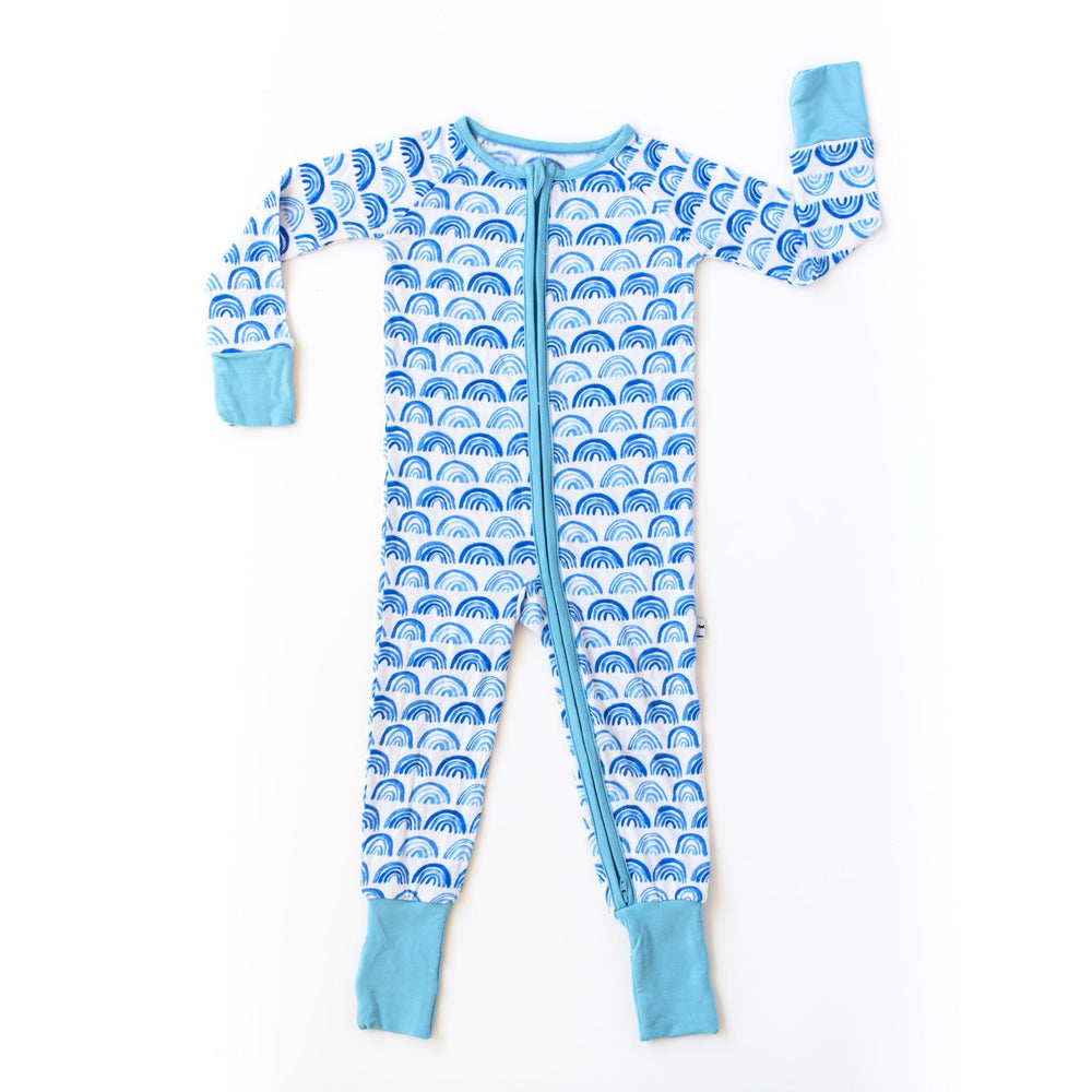 Click to see full screen - Flat lay photo of blue rainbow printed zip up romper. This print sits on a white background with shades of blue rainbows and sky blue trim details.