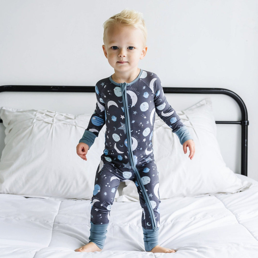 Click to see full screen - Image of little boy wearing a long sleeve zip up romper in Blue To the Moon & Back print. This print features blue and gray moons, stars, and planets on a charcoal background with dusty gray trim.