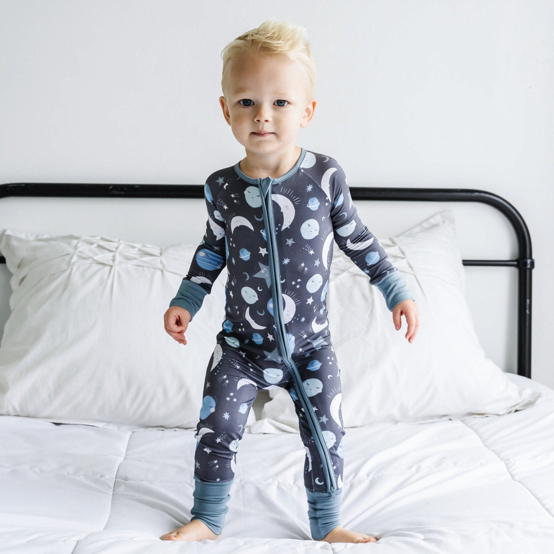 Image of little boy wearing a long sleeve zip up romper in Blue To the Moon & Back print. This print features blue and gray moons, stars, and planets on a charcoal background with dusty gray trim.