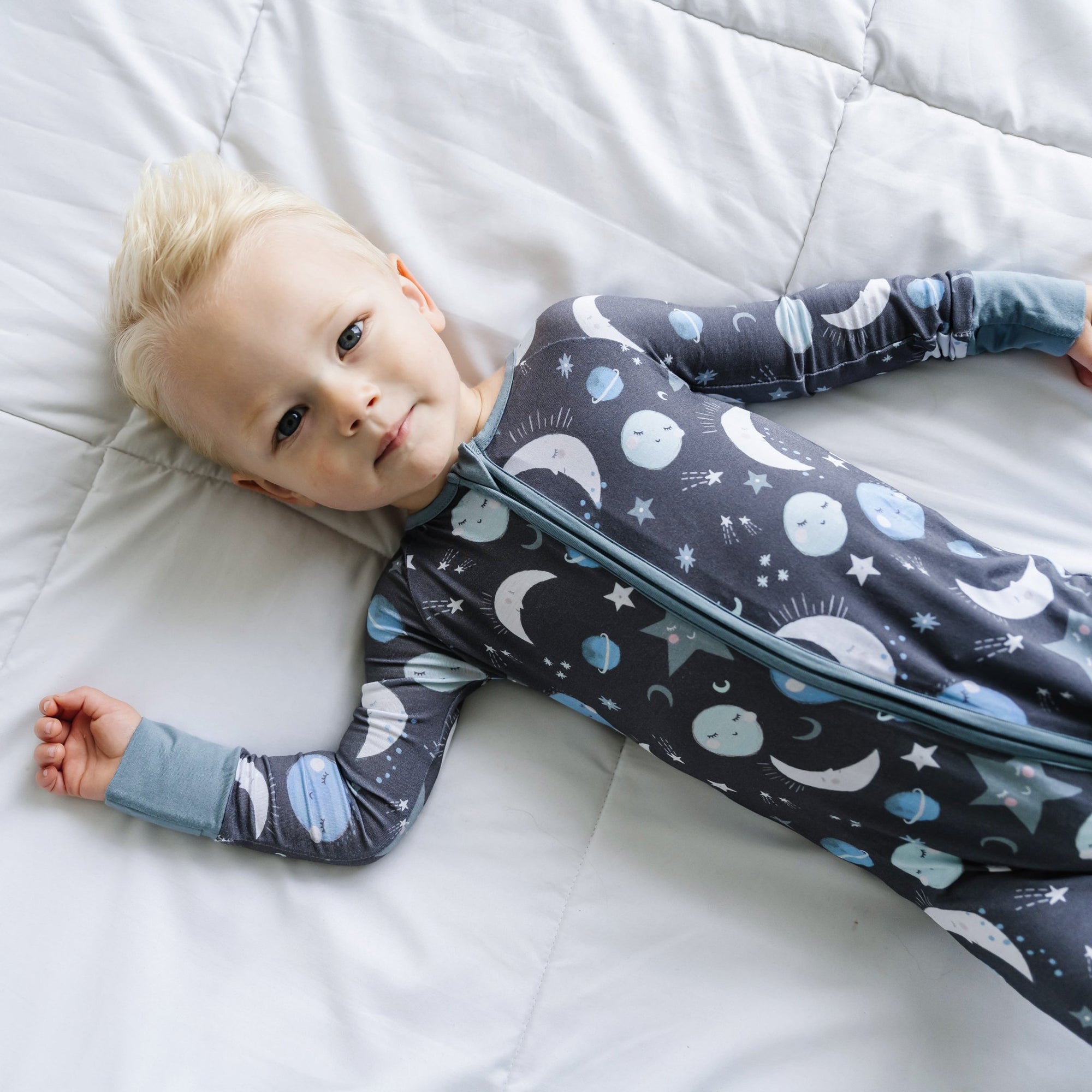 Image of little boy wearing a long sleeve zip up romper in Blue To the Moon & Back print. This print features blue and gray moons, stars, and planets on a charcoal background with dusty gray trim.