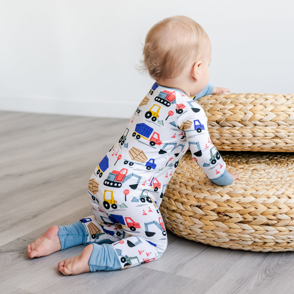 Click to see full screen - Image of baby boy wearing zip up romper in construction print. This print features utility trucks and tractors on a white background with sky blue trim accents.