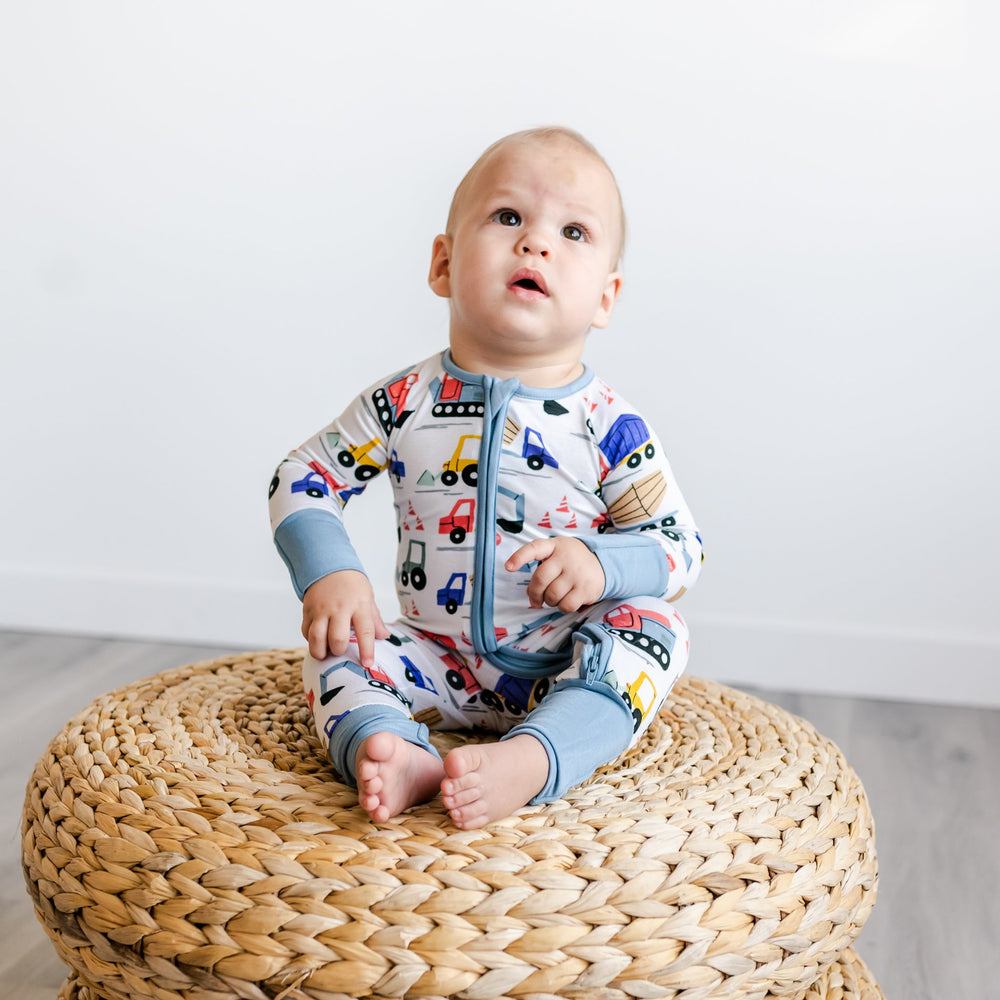 Image of baby boy sitting on rattan pouf. He is shown wearing a zip up romper in construction print. This print features utility trucks and tractors on a white background with sky blue trim accents.