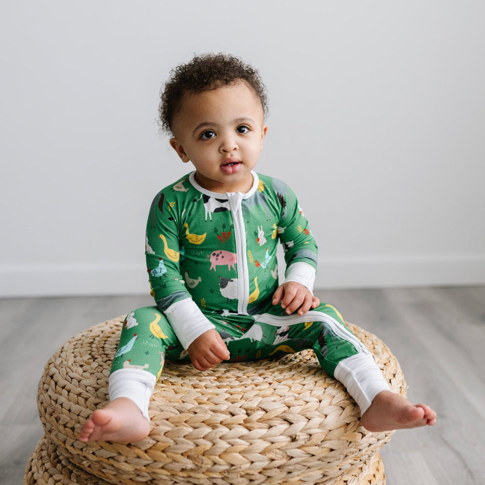 Image of toddler boy sitting on a stack of rattan poufs. He is shown wearing a zip up romper in the green farm animals print. This print includes a green background with white trim details. The farm animals featured on this print include cows, pigs, ducks