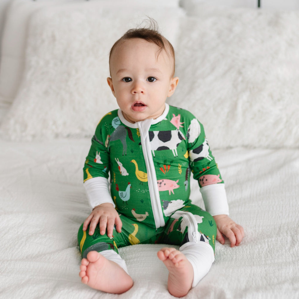 Image of infant boy sitting on a bed. He is shown wearing a zip up romper in the green farm animals print. This print includes a green background with white trim details. The farm animals featured on this print include cows, pigs, ducks, sheep, pigs, chic
