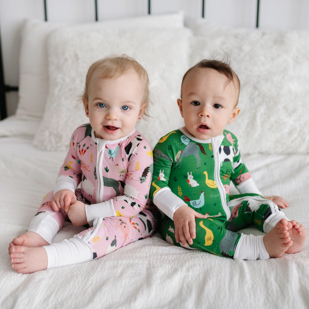 Image of infant boy and girl sitting next to each other on a bed. They are both shown wearing farm animal printed zip up rompers, with the boy in the green zip up romper and girl in pink. The farm animals featured on this print include cows, pigs, ducks, 