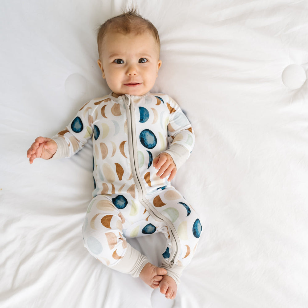 Image of a baby in a long sleeve zipper pajama romper in Luna Neutral print. This print features phases of the moon in the sweetest shades of creams, tans, and navy watercolor in an all over repeat pattern.