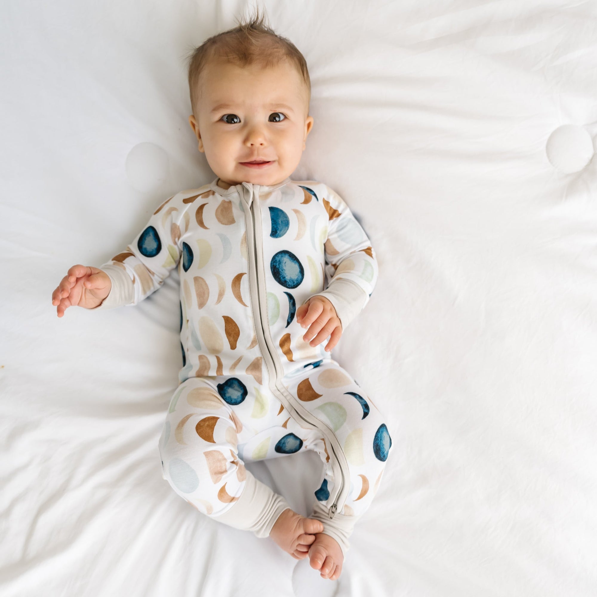 Image of a baby in a long sleeve zipper pajama romper in Luna Neutral print. This print features phases of the moon in the sweetest shades of creams, tans, and navy watercolor in an all over repeat pattern.