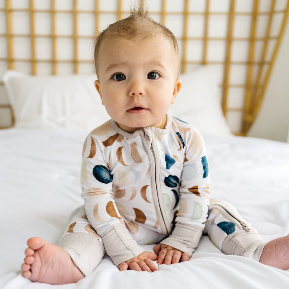 Image of a baby in long sleeve zipper romper pajamas in Luna Neutral print. This print features phases of the moon in the sweetest shades of creams, tans, and navy watercolor in an all over repeat pattern.