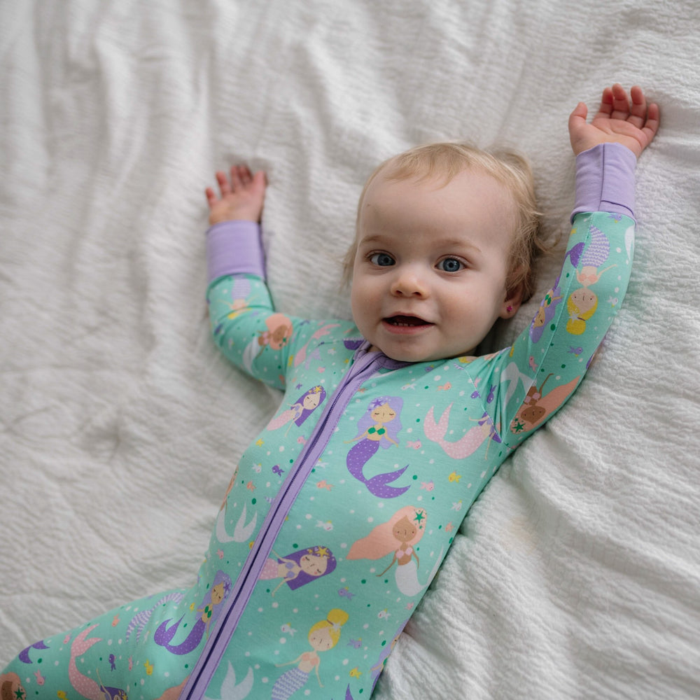 Image of infant girl wearing a mermaid printed zip up romper. This print includes multi-colored mermaids and fish that are featured on an aqua background with a purple trim. 