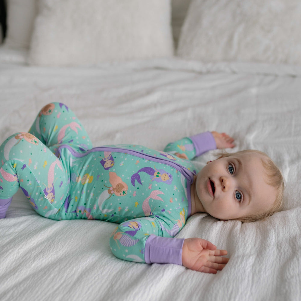 Image of infant girl wearing a mermaid printed zip up romper. This print includes multi-colored mermaids and fish that are featured on an aqua background with a purple trim.