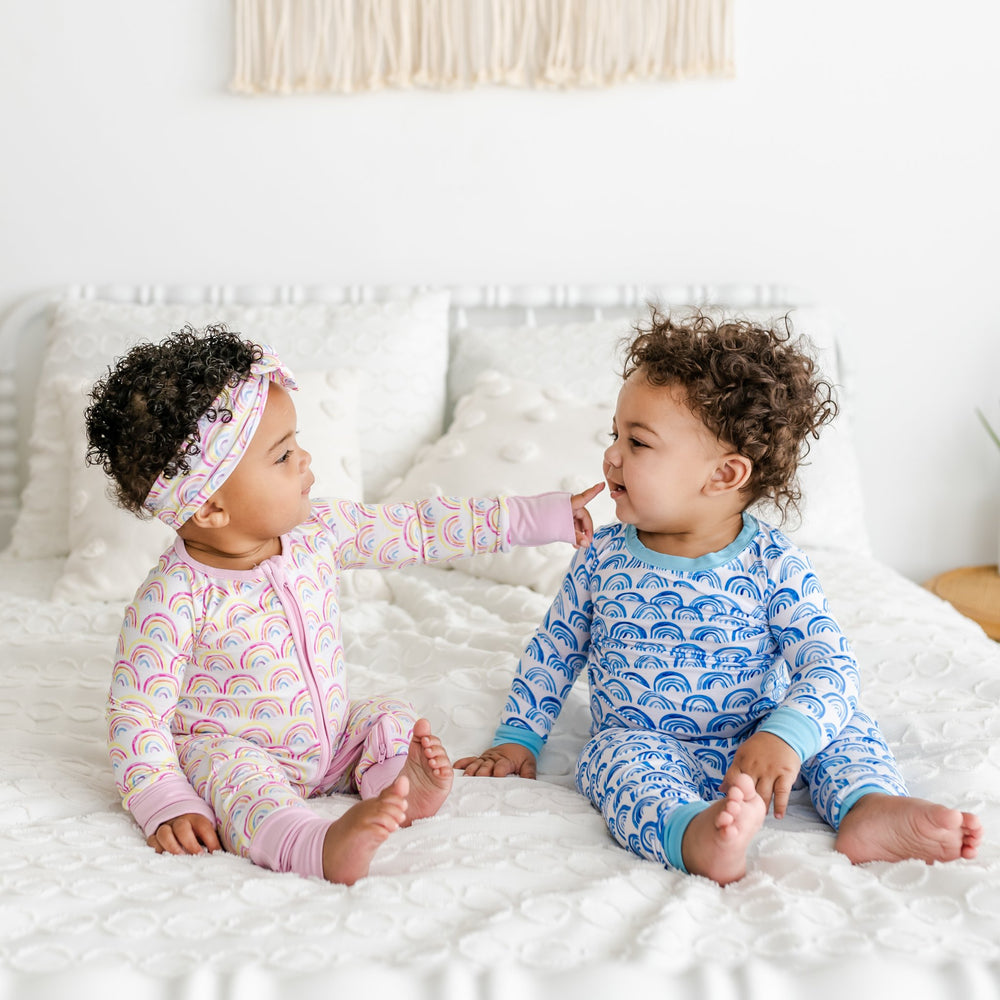 Click to see full screen - Image of infant boy and girl wearing matching rainbow printed pajamas. Infant girl is wearing rainbow printed zip up romper in pink with matching bow headband, and infant boy is wearing rainbow printed two-piece pajama set in blue.