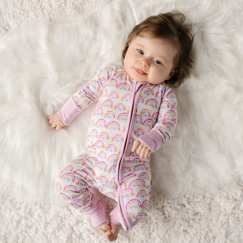 Click to see full screen - Image of infant girl wearing rainbow printed zip up romper with matching bow headband. This print features multicolored rainbows that sit upon a white background with light pink accent trim.