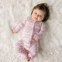 Image of infant girl wearing rainbow printed zip up romper with matching bow headband. This print features multicolored rainbows that sit upon a white background with light pink accent trim.