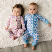 Image of infant boy and girl wearing matching rainbow printed pajamas. Infant girl is shown wearing zip up romper in pink and boy is wearing zip up romper in blue. 
