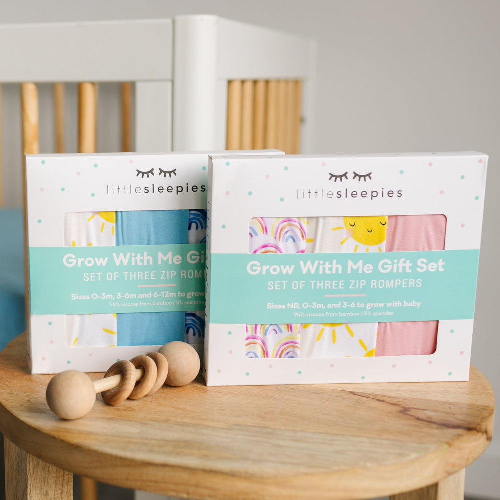 Click to see full screen - Image of two Grow With Me gift boxes photographed together on a wooden table. The gift box on the left contains a set of 3 zip up rompers in sunshine, solid sky blue, and blue rainbows. The gift box pictured on the right contains a set of 3 zip up rompers in pastel rainbows, sunshine, and bubblegum pink