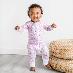 Image of infant girl wearing breakfast printed zip up romper. This print has a light pink background with white trim accents and the breakfast foods featured on this print include sunny side up eggs, toast, and milk.