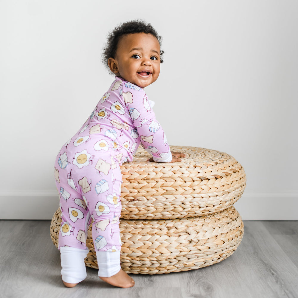 Image of infant girl wearing breakfast printed zip up romper. This print has a light pink background with white trim accents and the breakfast foods featured on this print include sunny side up eggs, toast, and milk.