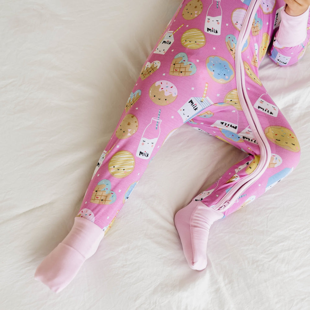 Zoomed in image of toddler girl wearing a zip up romper in cookies and milk print. The image shows the cuffs on the zip up romper folded over her feet to cover them. This print features milk cartons, colorful sprinkled cookies, and chocolate chip cookies 
