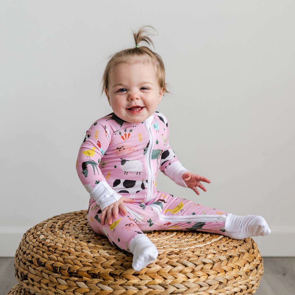 Click to see full screen - Child sitting on woven pouf wearing Pink Farm Animals zippy with feet cuffs folded over