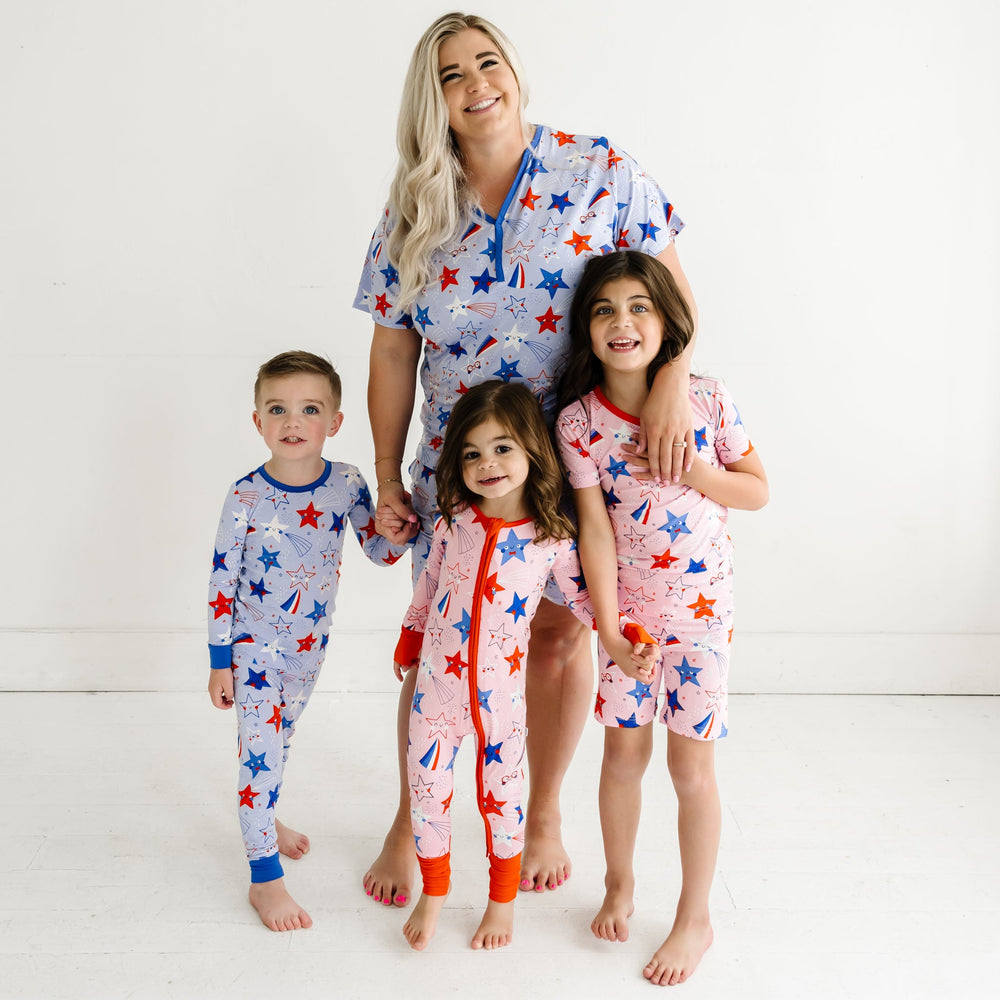 Mom and her three children posing together wearing coordinating Stars and Stripes printed pajamas. Mom is wearing a Blue Stars and Stripes printed women's pajama top and matching women's pajama shorts. One child is wearing a Blue Stars and Stripes printed two piece pajama set and two children are wearing Pink Stars and Stripes printed two piece short sleeve and shorts pajama set and zippy
