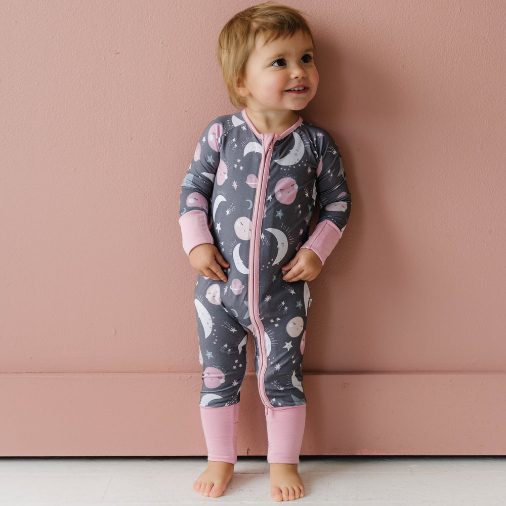 Image of toddler girl wearing a long sleeve zip up romper in pink to the moon and back print. This print features pink and gray moons, stars, and planets on a charcoal background with a matching pink trim. 