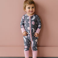 Image of toddler girl wearing a long sleeve zip up romper in pink to the moon and back print. This print features pink and gray moons, stars, and planets on a charcoal background with a matching pink trim. 