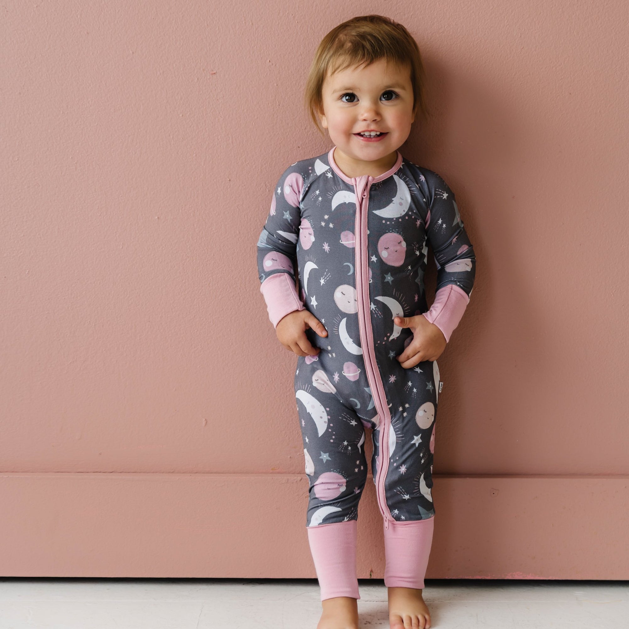 Image of toddler girl wearing a long sleeve zip up romper in pink to the moon and back print. This print features pink and gray moons, stars, and planets on a charcoal background with a matching pink trim. 